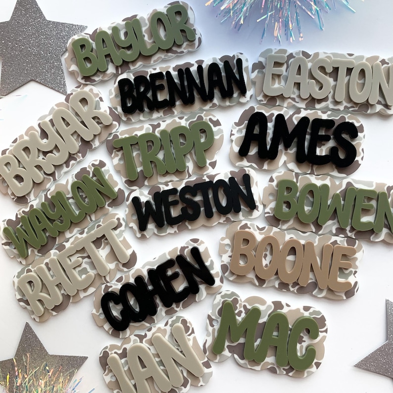 May include: A collection of personalized name tags with a camouflage pattern background. The names are in various colors, including green, black, and tan. The names are in a bold, rounded font.