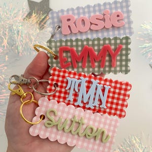 May include: Four rectangular keychains with scalloped edges and gingham patterns in light blue, sage green, red, and light pink. Each features raised, three-dimensional lettering in pink, red, light blue, and sage green spelling Rosie, EMMY, TMN, and Sutton.