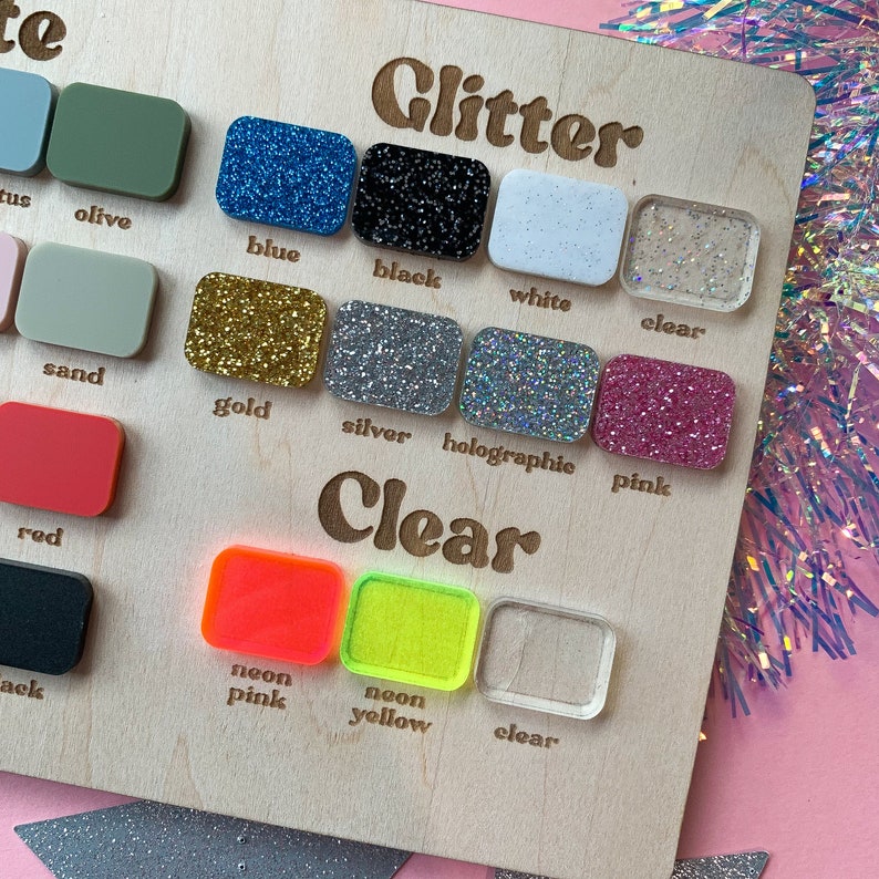 May include: A wooden display board featuring a variety of small, rectangular samples in different colors and finishes. The samples include glitter and clear options, with colors like blue, black, gold, silver, pink, and neon. The word "Glitter" and "Clear" are written on the board.