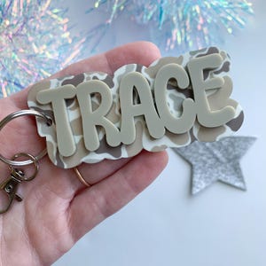 May include: A custom keychain with the name "TRACE" in light gray, layered over a camouflage pattern in shades of beige and brown. The keychain includes a silver keyring and clasp for easy attachment.
