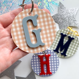 May include: Round keychains with a gingham pattern and a letter in the center. The letters are in blue, red, and navy. The background is a light brown, blue, and green gingham pattern. A silver star is in the background.