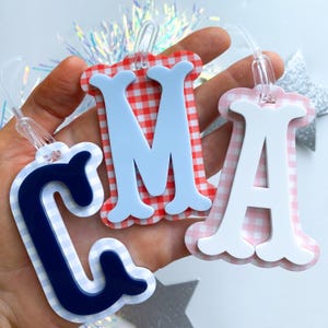 May include: Three luggage tags with the letters "C", "M", and "A". The tags have a gingham pattern in red and white, navy blue and white, and pink and white. The letters are light blue, white, and white, respectively. Each tag has a clear plastic loop.