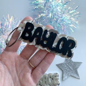 May include: A custom luggage tag with the name "BAYLOR" in black lettering over a camouflage pattern. The tag is attached to a silver wire loop. A gold ring is visible on a finger holding the tag.