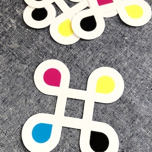 May include: White paper cutouts in a four-leaf clover shape, each with a colored teardrop design in cyan, magenta, yellow, and black. The cutouts are scattered on a textured blue surface.