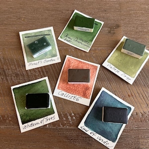 May include: A collection of watercolour paint samples on small cards. Each card displays a different colour swatch with the paint name handwritten below. Colours include green, brown, and blue. The cards are arranged on a wooden surface.