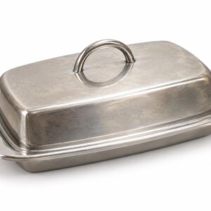May include: A rectangular stainless steel butter dish with a matching lid. The base features integrated side handles, while the lid is topped with a curved, arched handle. The metal surface has a smooth, brushed finish with a subtle metallic sheen.