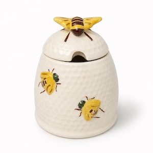 May include: A cream-colored ceramic honey pot with a textured honeycomb pattern. The lid features a sculpted yellow and brown bee handle. Two additional yellow and brown bees are painted on the body of the pot, which includes a small notch for a dipper.