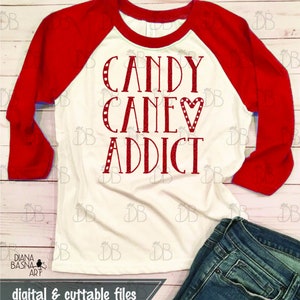 Peut inclure: Un t-shirt de baseball blanc et rouge avec le texte "CANDY CANE♡ ADDICT" en lettres scintillantes rouges. Le t-shirt est pos&eacute; sur une surface en bois avec un jean bleu.