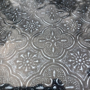May include: A close-up of a silver metallic surface with a repeating embossed pattern of four-leaf clover shapes. The surface has a subtle sheen and a slightly textured feel.