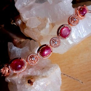 May include: A rose gold bracelet featuring alternating oval cabochon-cut red star rubies and circular links. Each ruby is surrounded by a halo of small white stones, while the circular links contain a central white stone within a filigree pattern.