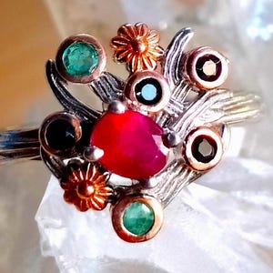 Peut inclure: Une bague en argent avec une pierre centrale ovale rouge entourée de petites pierres rondes vertes, bleu foncé et noires, agrémentée de deux petites fleurs en métal cuivré. L'anneau présente un aspect texturé rappelant une branche.
