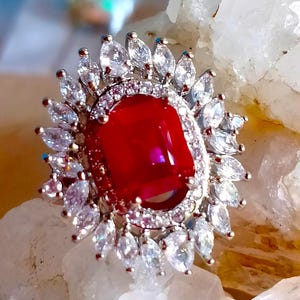 Könnte beinhalten: Ein silberfarbener Ring mit einem großen, rechteckigen, leuchtend roten Mittelstein. Der Mittelstein ist von einem Kranz aus kleinen, klaren, marquise-geschliffenen Steinen in einem strahlenförmigen Muster auf einer Metallschiene umgeben.