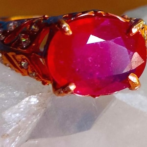 Peut inclure: Une bague dorée ornée d'une grande pierre précieuse rouge vif de taille ovale, maintenue par quatre griffes. L'anneau est décoré d'une série de petites pierres claires de taille ronde incrustées sur les côtés dans une monture métallique texturée.