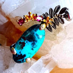 May include: A pendant featuring an oval turquoise stone with black matrix, accented by a dark metal fern-like leaf structure, small gold-toned flowers, and embedded red and yellow gemstones, all secured by metal prongs in a decorative arrangement.