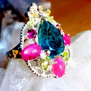 Peut inclure: Une bague argentée ornée d'une pierre bleue centrale en forme de poire, entourée de pierres rose vif ovales, de petits accents rose vif ronds et de minuscules gemmes vert clair, le tout serti dans une monture ajourée aux motifs travaillés.