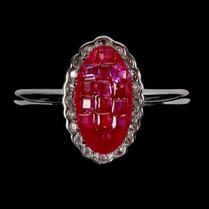 Puede incluir: Un anillo de plata con una pieza central ovalada compuesta por múltiples rubíes rojos de corte cuadrado dispuestos en cuadrícula, rodeada por un borde festoneado de pequeñas piedras claras en una banda de metal plateado.