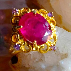 May include: A gold ring featuring a large, oval-cut, vibrant pinkish-red center stone held by prongs. The setting is crafted with intricate, swirling gold metalwork and accented by four small, round, light purple stones at the corners.