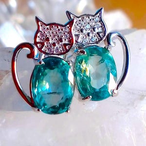Puede incluir: Un par de pendientes de plata con forma de gato con piedras preciosas verdes. Los gatos se miran y tienen pequeñas piedras blancas en la cabeza.