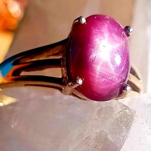 Può includere: Un anello in oro con una grande pietra preziosa rubino stellato di forma ovale e taglio a cabochon. La pietra color magenta intenso mostra un distinto effetto a stella a sei punte sulla superficie, fissata da quattro griffe metalliche.