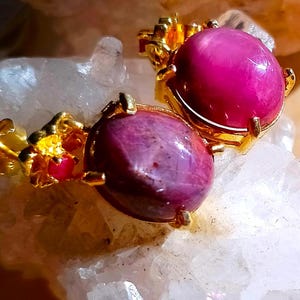 May include: A pair of gold stud earrings featuring large, oval-shaped, deep pink cabochon gemstones. Each earring is accented with a smaller, round, bright pink stone set within a textured gold floral-inspired metal frame surrounding the main gem.