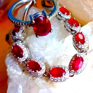 Op de afbeelding: Een zilverkleurige ring en een bijpassende armband met ovaal geslepen, levendig rode stenen omringd door kleine, helderwitte steentjes. De sieraden zijn in een cirkelvorm gerangschikt, waarbij de gefacetteerde rode edelstenen en metalen zettingen zichtbaar zijn.