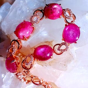 May include: A rose gold bracelet featuring five oval-shaped, deep pink cabochon gemstones. Each gemstone is separated by a small, teardrop-shaped link encrusted with tiny clear stones. The bracelet has a delicate chain and a secure metal clasp.