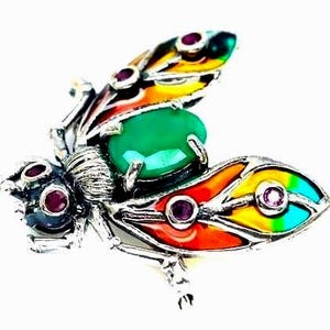 Può includere: Una spilla a forma di insetto color argento con una grande pietra centrale ovale verde, piccole pietre rotonde rosse sulla testa e sulle ali, e sezioni alari in smalto traslucido in vivaci tonalità arancione, giallo e verde acqua metallizzato.
