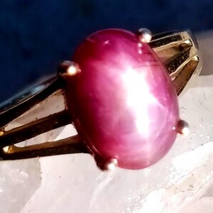 May include: A gold ring with a large, oval, purplish-red gemstone. The gemstone is secured by four small prongs. The ring's band has a simple, split design.