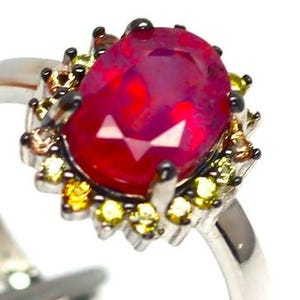 Può includere: Un anello d'argento con una grande pietra preziosa rosso scuro a taglio ovale come elemento centrale. La pietra preziosa è circondata da pietre di accento gialle e marroni più piccole. L'anello ha un'incastonatura dettagliata e ornata.