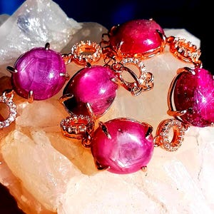 May include: A rose gold bracelet featuring four large, oval-shaped, deep pink cabochon rubies held in prong settings. The links between the rubies consist of small, circular rose gold frames encrusted with tiny, clear, sparkling white stones.