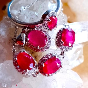 May include: A silver-toned jewellery set consisting of a ring, a pendant, and a pair of earrings. Each piece features a vibrant oval-cut red gemstone surrounded by a halo of small clear stones. The metal settings are polished and reflective.