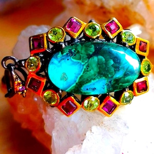 May include: A pendant featuring a large, oval-shaped, teal and green patterned stone at the centre. The stone is surrounded by a decorative border of alternating square-cut pink gemstones and round, light green faceted stones in gold settings.