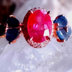 May include: A ring featuring a large, oval-cut vibrant red gemstone as the centerpiece, surrounded by a halo of small clear stones. The band is rose-colored metal, accented on both sides by clusters of three deep blue, pear-shaped gemstones.