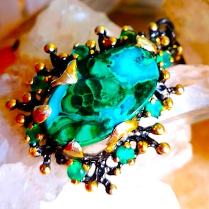 May include: An oval malachite pendant featuring a dark, textured metal setting with gold-toned accents and small, round green stones embedded along the perimeter. The central malachite stone displays deep green and teal banding patterns.