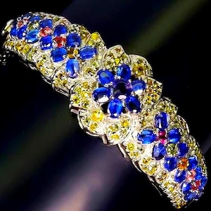May include: A silver-toned bracelet featuring a series of floral motifs composed of oval-cut blue gemstones. The flowers are surrounded by clusters of small, round yellow and green gemstones, creating a vibrant, multi-colored botanical pattern.