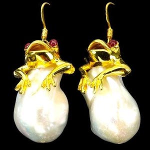 May include: A pair of gold-coloured frog earrings with red gemstone eyes. The frogs are perched on large, white, teardrop-shaped pearls. The earrings have gold-coloured hooks against a black background.