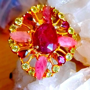 May include: A gold ring featuring a large central pear-shaped deep red gemstone, surrounded by smaller pink leaf-shaped stones, dark red teardrop-shaped stones, and tiny round light green stones arranged in a circular, openwork metal setting.