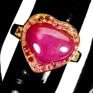 Puede incluir: Un anillo de oro con una gema rosa grande, lisa y en forma de corazón en el centro. El corazón está rodeado por un halo de piedras más pequeñas, redondas, facetadas en tonos rosa y rojo, engastadas en un marco de oro que sigue por los lados.