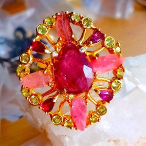 Puede incluir: Un anillo dorado con un gran rubí facetado en forma de lágrima como pieza central. El anillo está adornado con varias piedras preciosas en tonos rosa, rojo y amarillo verdoso, engastadas en un diseño calado detallado. El anillo se exhibe sobre una superficie blanca.