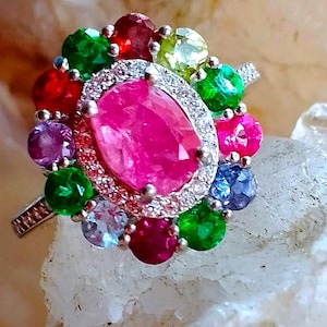 Op de afbeelding: Een zilveren ring met een grote ovaal geslepen roze middensteen, omringd door een halo van kleine heldere steentjes. De buitenrand bestaat uit meerdere rond geslepen edelstenen in levendige tinten rood, groen, paars, blauw en lichtgeel.