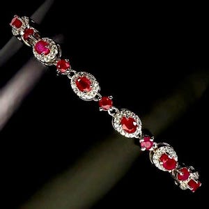 Puede incluir: Una pulsera de tono plateado que presenta una serie de rubíes rojos de forma ovalada, cada uno rodeado por un halo de pequeñas piedras blancas. El diseño alterna entre engastes ovalados más grandes y acentos de rubíes redondos más pequeños.