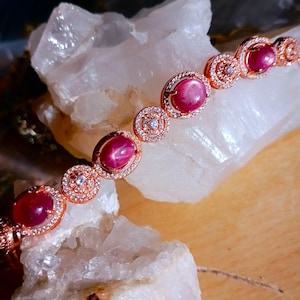 Puede incluir: Un brazalete de oro rosa con piedras preciosas de rubí ovaladas con forma de estrella. El brazalete presenta una serie de eslabones circulares y ovalados, cada uno adornado con pequeñas gemas transparentes. Se muestra sobre una superficie cristalina blanca.