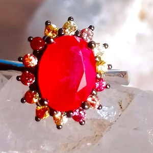 Può includere: Anello d'argento con una grande gemma rossa sfaccettata al centro, circondata da gemme più piccole colorate in rosso, arancione, giallo e rosa. Il design dell'anello ricorda un'esplosione solare.