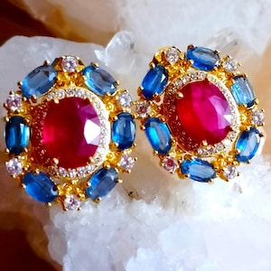 Puede incluir: Un par de pendientes dorados con una gran piedra central roja de corte ovalado rodeada por un halo de pequeñas piedras blancas, seguido de un anillo exterior de piedras azules ovaladas y pequeñas piedras blancas alternadas.