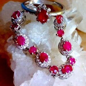 Op de afbeelding: Een zilverkleurige ring en een bijpassende armband met ovaal geslepen, levendig rode edelstenen omringd door kleine heldere steentjes. De sieraden zijn in een cirkelvorm gerangschikt, waarbij de gepolijste metalen zettingen en de helderrode kleur goed zichtbaar zijn.
