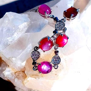 May include: A silver-toned pendant featuring multiple oval-shaped cabochon gemstones in shades of pink and deep red, accented with small clear pavé-set stones. The jewelry piece has a dangling, multi-section design with prong settings for each stone.