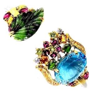 Könnte beinhalten: Ein Goldring mit einem großen ovalen blauen Topas, umgeben von einer Ansammlung kleiner runder, mehrfarbiger Edelsteine wie Granat, Peridot und Citrin, zusammen mit einer separaten Goldbrosche in Form eines geschnitzten grünen Blattes.