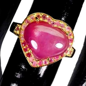 Peut inclure: Une bague en or avec une grande pierre précieuse rose en forme de cœur lisse au centre. Le cœur est entouré d'un halo de petites pierres rondes roses et rouges serties dans une monture en or. L'anneau de la bague est également en or.