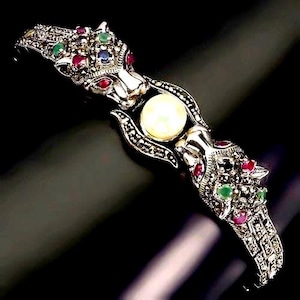 Könnte beinhalten: Ein silberfarbenes Gliederarmband mit zwei einander zugewandten Pantherköpfen. Die Pantherköpfe sind mit kleinen roten, grünen und blauen Edelsteinen sowie dunklen Akzenten besetzt, mit einer einzelnen runden weißen Perle in der Mitte.