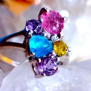 Könnte beinhalten: Ein Silberring mit einem Cluster aus farbigen Edelsteinen. Der Ring zeigt eine lebendige Anordnung aus rosa, blauen, gelben und lila Edelsteinen. Die Edelsteine sind in einem Silberband gefasst, was einen auffälligen Kontrast erzeugt.